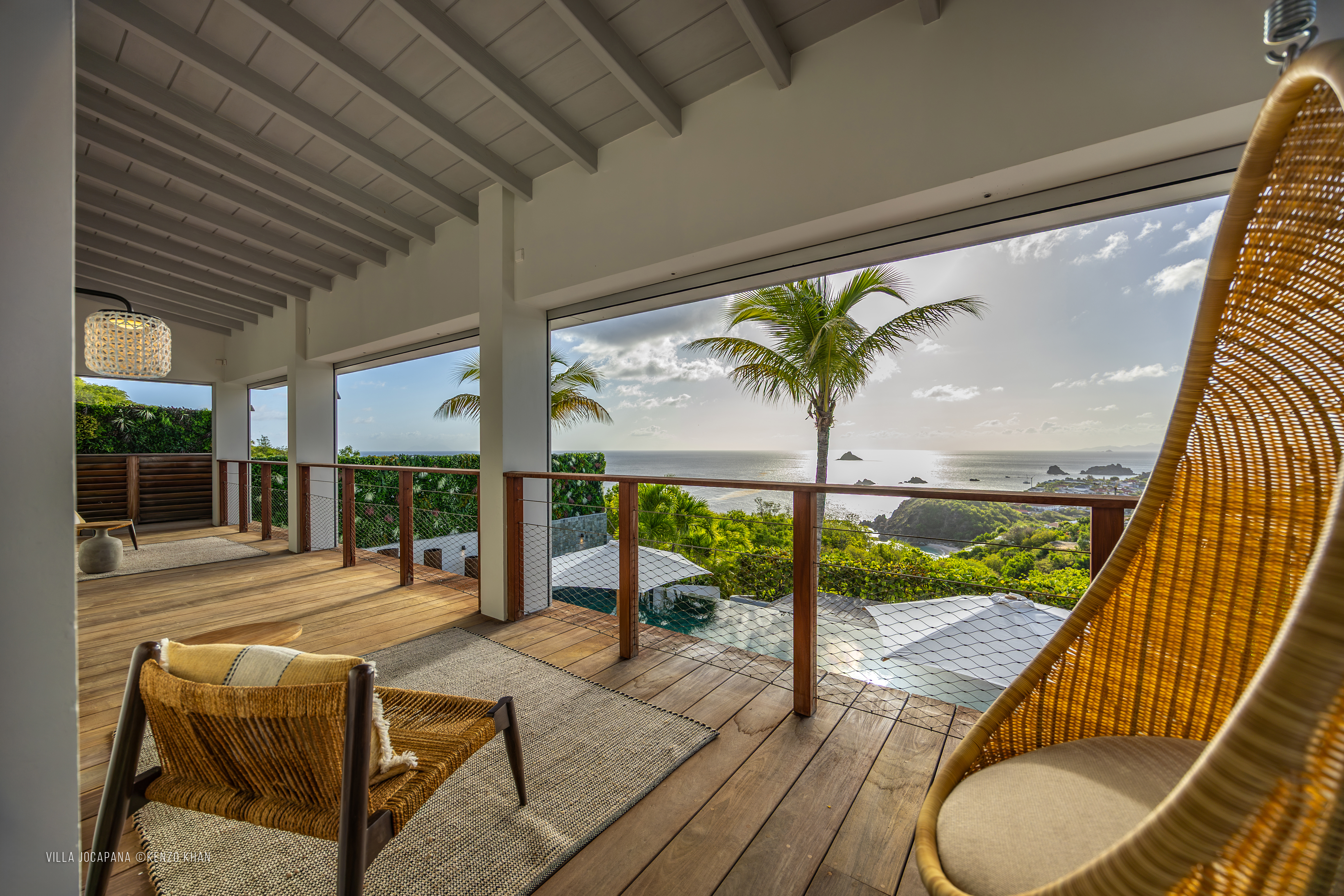 Covered terrace at Villa Jocapana with rattan hanging chair, lounge seating, and panoramic ocean views at sunset.