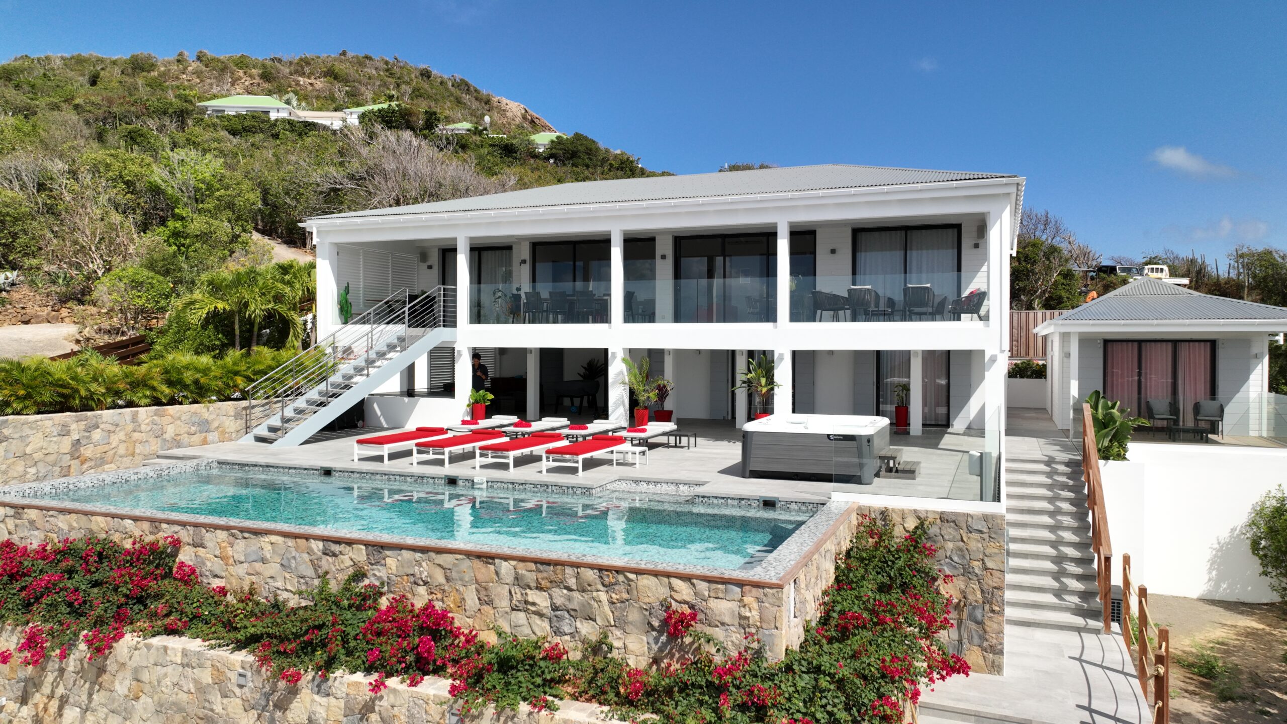 An aerial photo of a two-story modern villa and a smaller bungalow connected by a pathway. The pool terrace and lush landscaping are visible around the buildings.