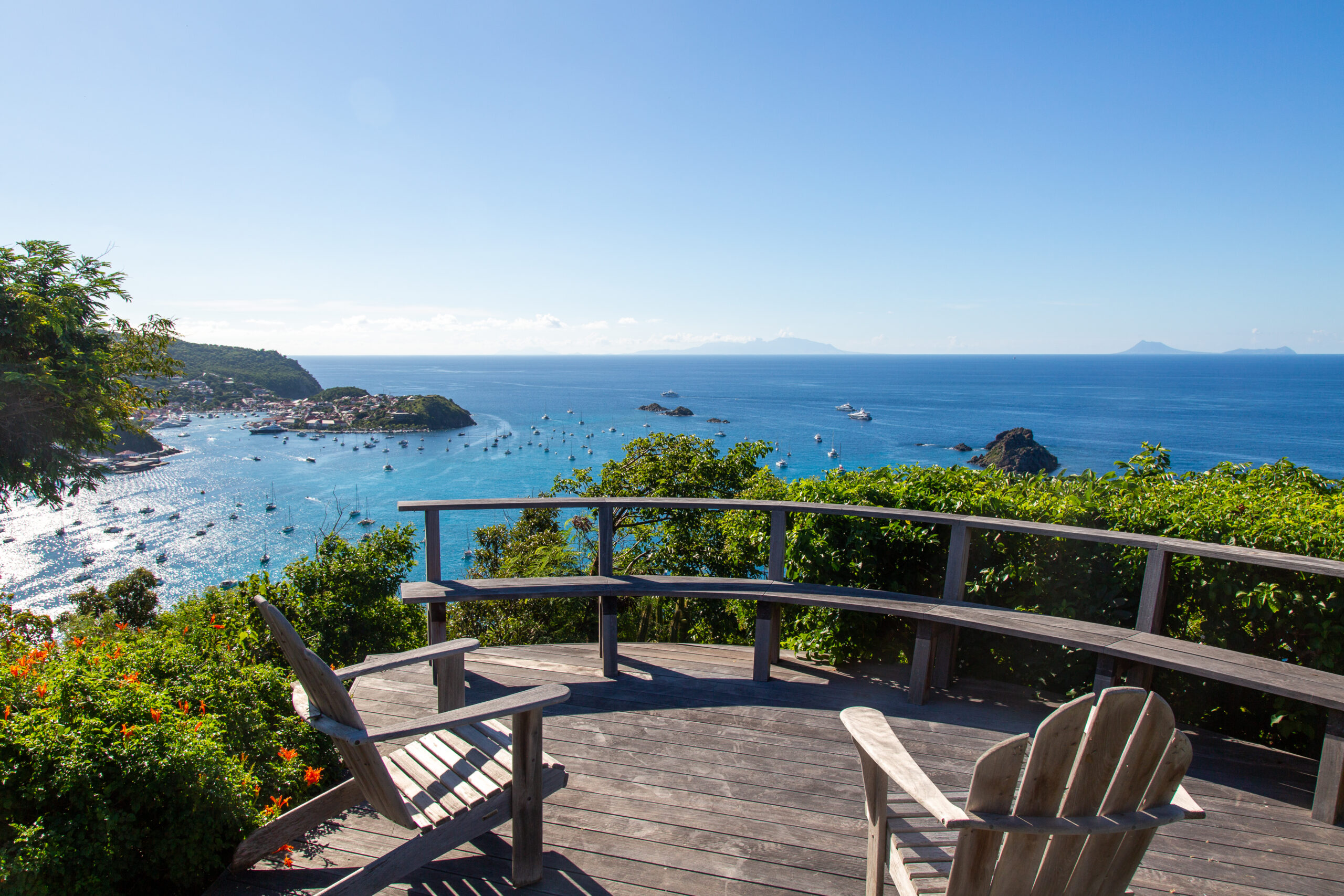 A beautiful wooden deck with Adirondack chairs and a curved bench, overlooking a sweeping bay and the ocean under a clear blue sky.