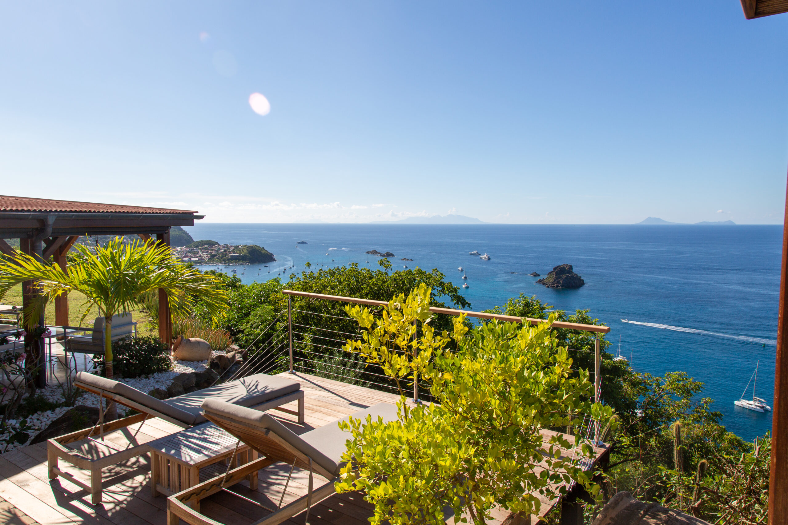 A wooden sun deck with two modern sun loungers overlooking a vibrant blue ocean and coastline.