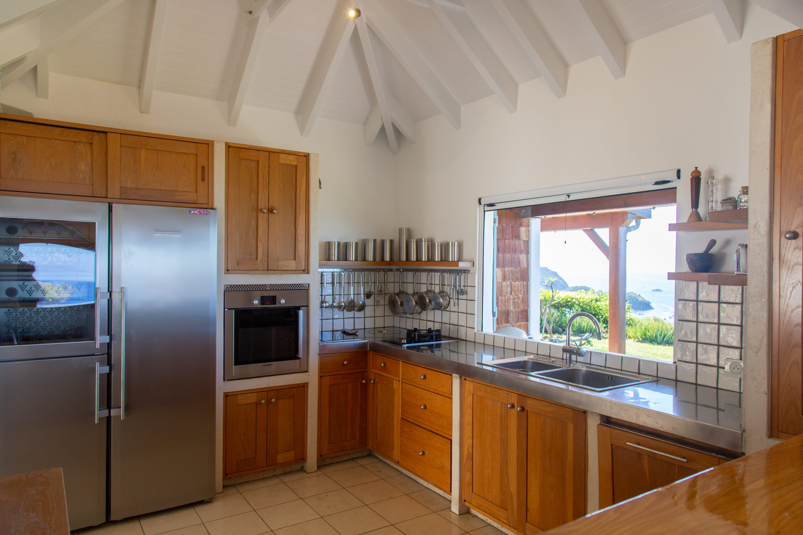 A modern and functional kitchen with wooden cabinets, stainless steel appliances, and a pass-through window with an ocean view.