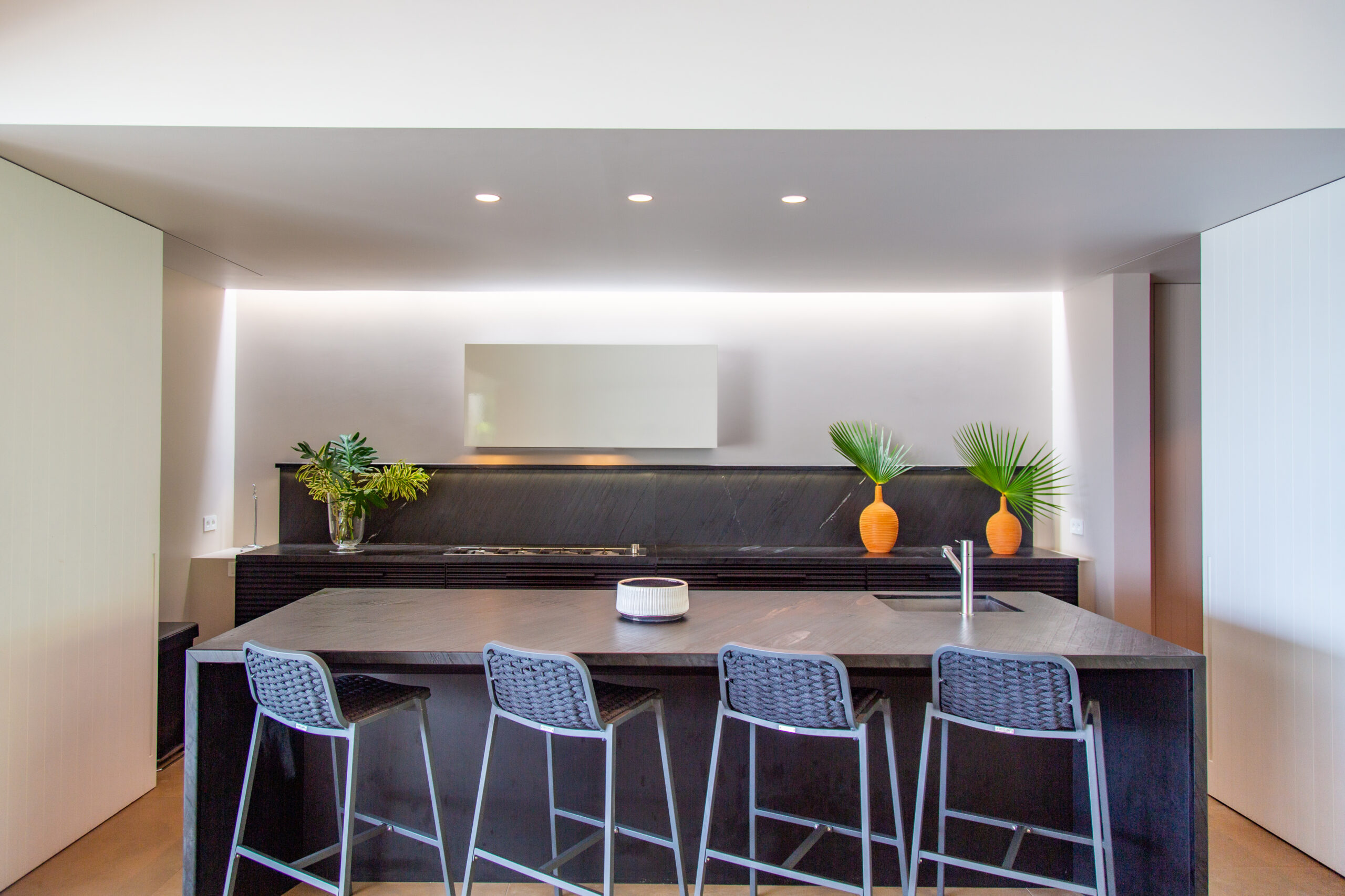 a modern kitchen with a large dark countertop island, four woven stools, and recessed lighting
