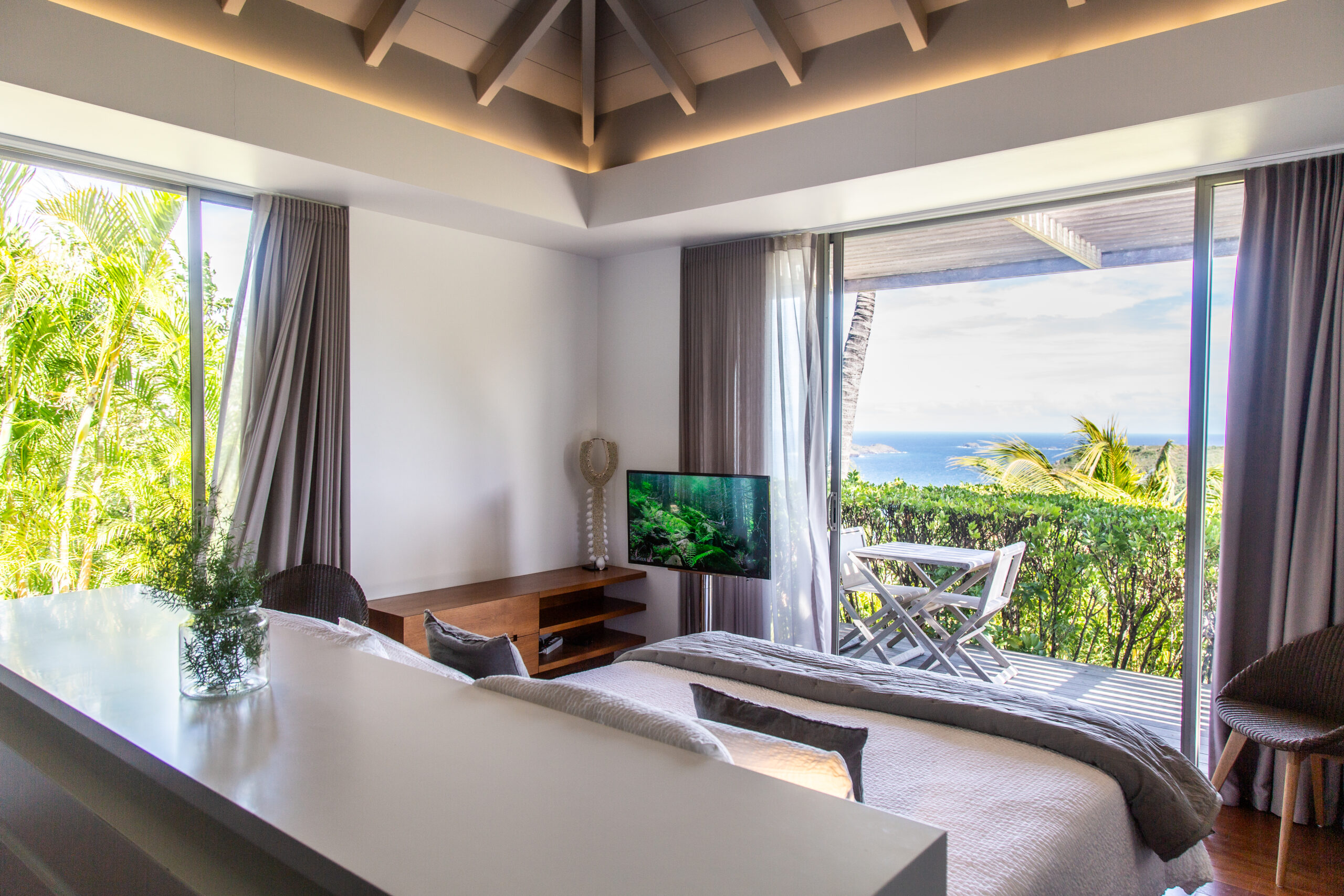 a bedroom with a king-size bed and a glass sliding door leading to a small outdoor terrace with a table, chairs, and an ocean view