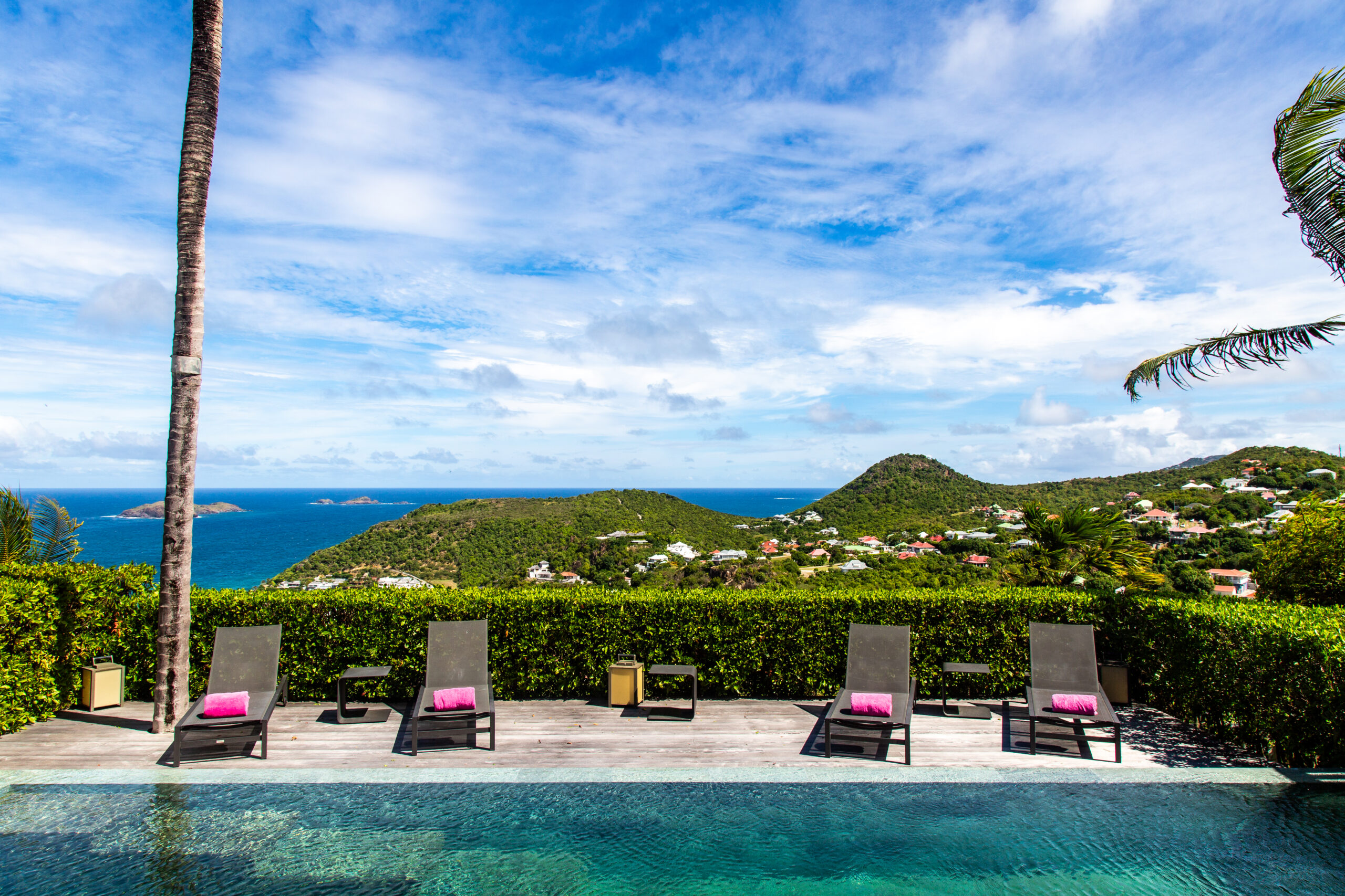 a wide shot of a poolside terrace with multiple lounge chairs facing a private pool and a magnificent panoramic view of the blue ocean and green hills